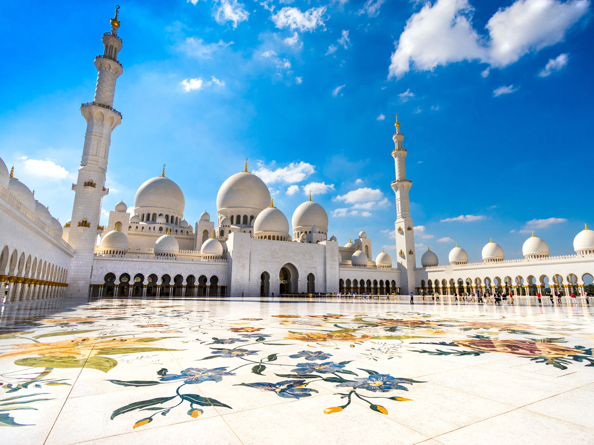 A large white building with domes and towers with Sheikh Zayed Mosque in the background  Description automatically generated